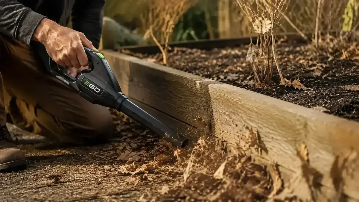 Person using a compact cordless leaf blower to clear leaves from a raised garden bed, crouching without straining